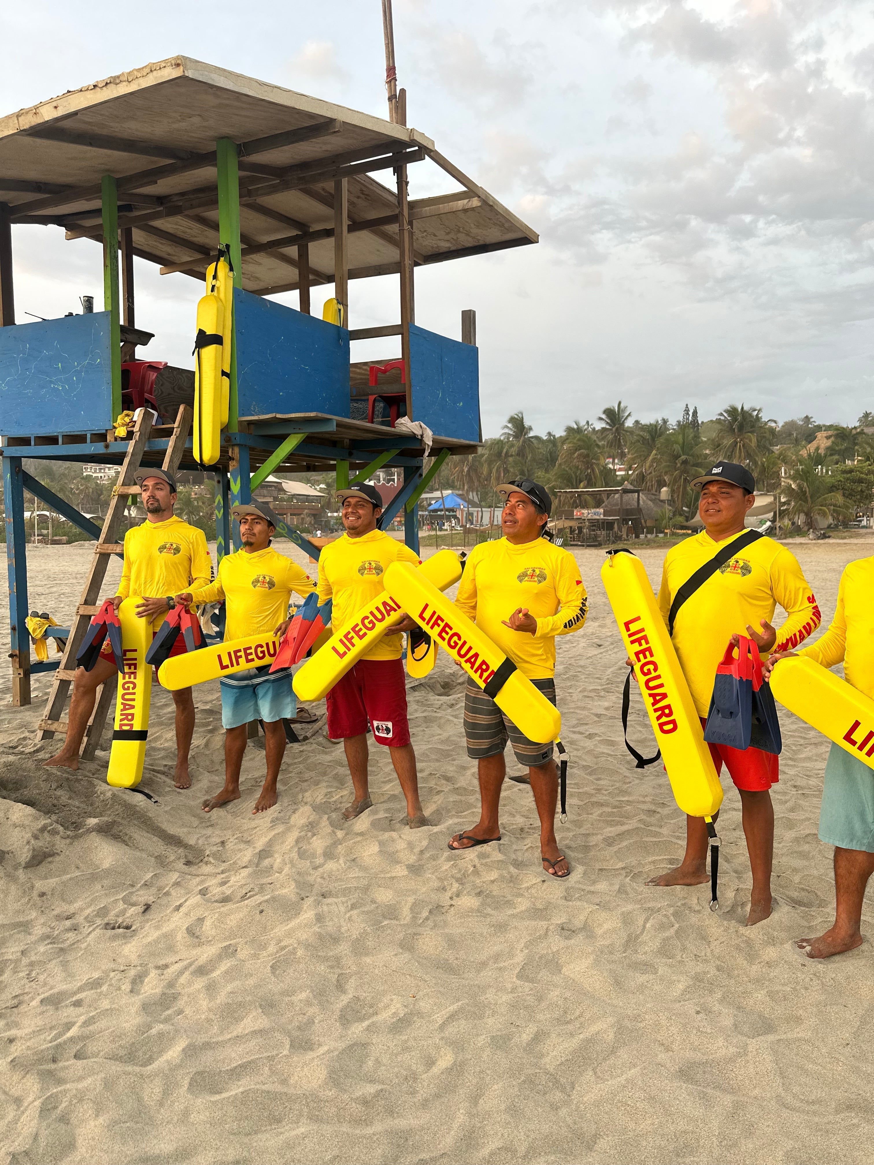 melin x BCMSF presents a team of lifeguards in yellow shirts holds rescue boards, standing in front of a beach tower on a sandy beach.