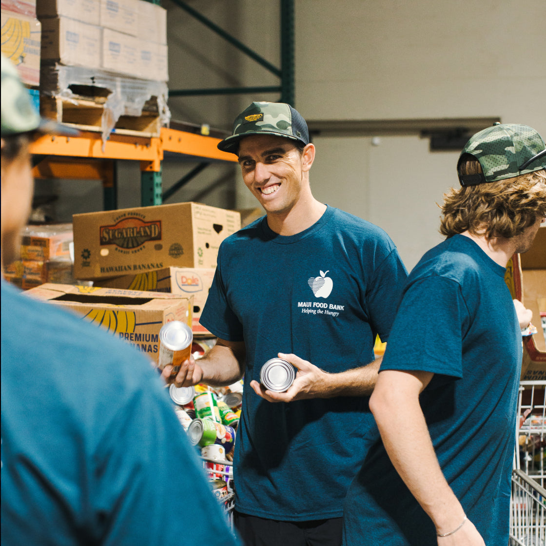 Two men in blue shirts with the Maui Food Bank logo, holding cans in a warehouse setting.