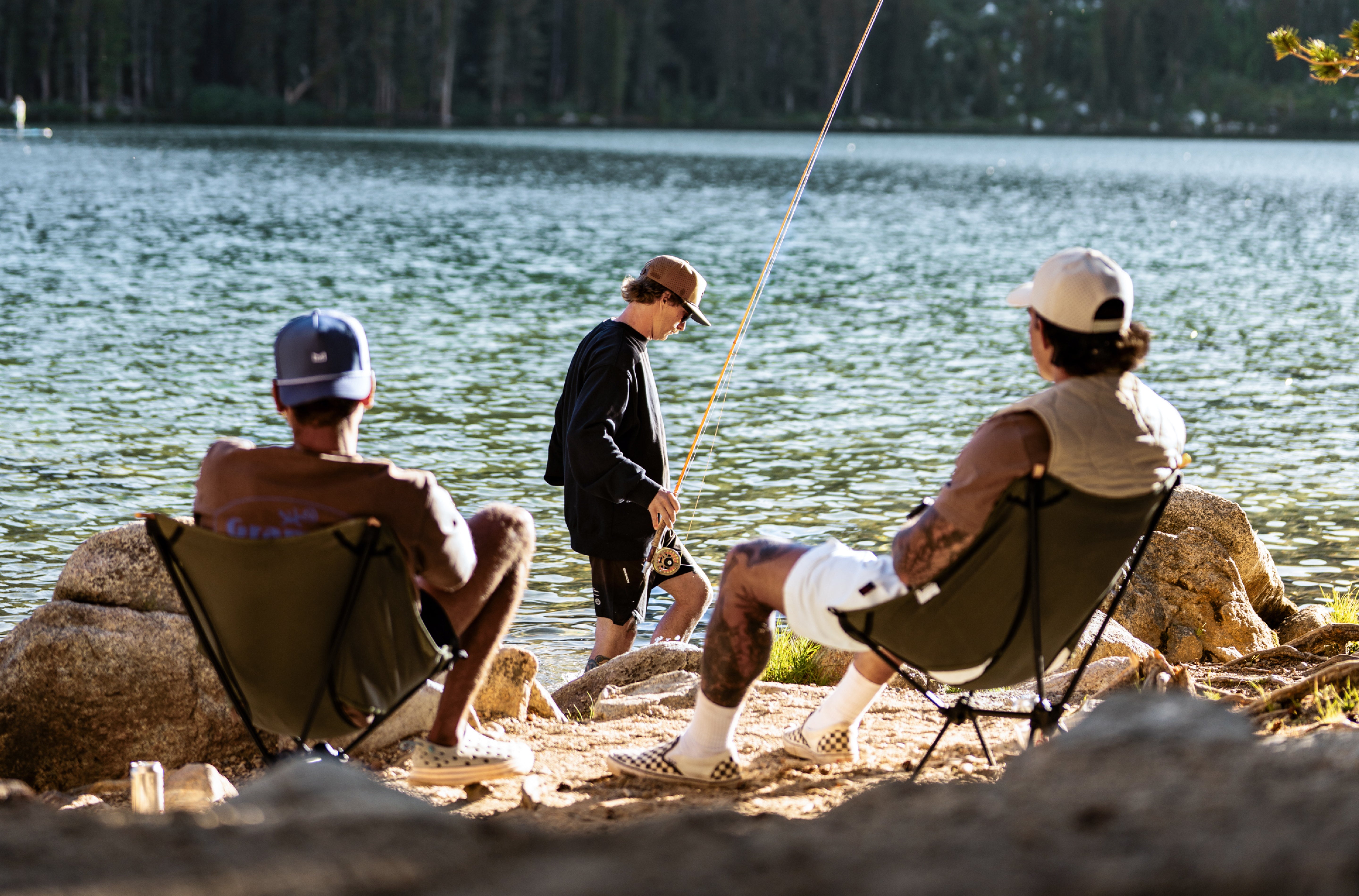 Three people sitting by a lake, two on chairs and one standing, with fishing equipment.