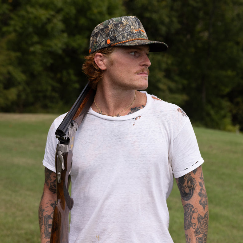 Man wearing the melin Coronado Brick Realtree Hydro holding a rifle in a grassy outdoor setting