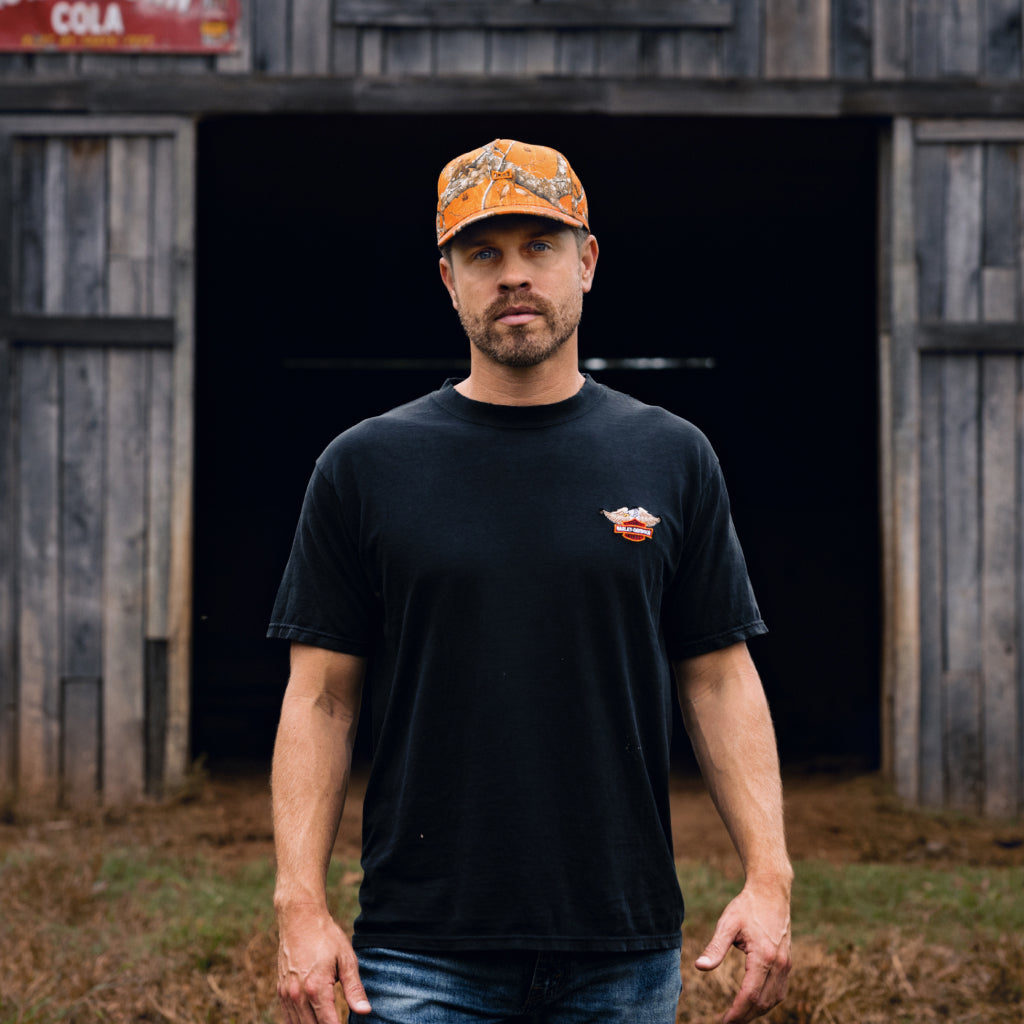 Man wearing a black t-shirt with a logo and the melin Odysea Realtree Hydro standing in front of a wooden building.